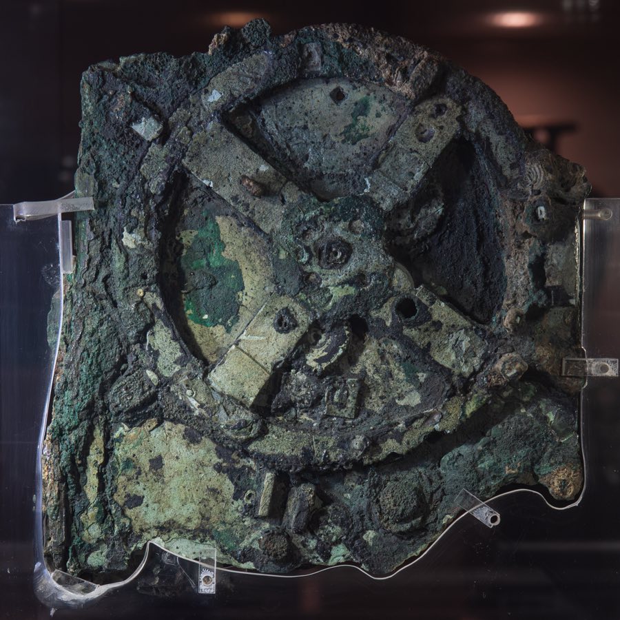 A roughly square slab of corroded bronze, mottled pale green over dark grey, held upright in a museum case by clear plastic supports, with a large wheel of four spokes and the rims of smaller toothed wheels showing through the crust.