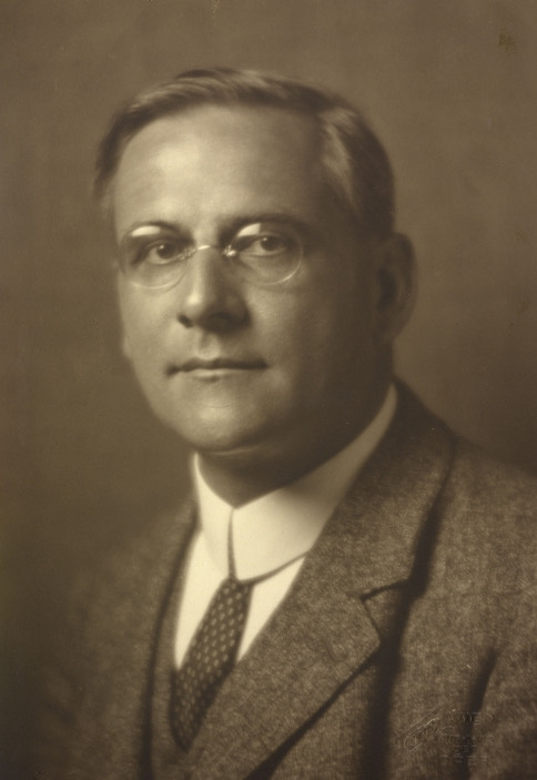A sepia studio portrait of a middle-aged man in round rimless spectacles, shown from the chest up in a tweed jacket, white shirt and patterned dark tie, turned slightly away from the camera against a plain backdrop.
