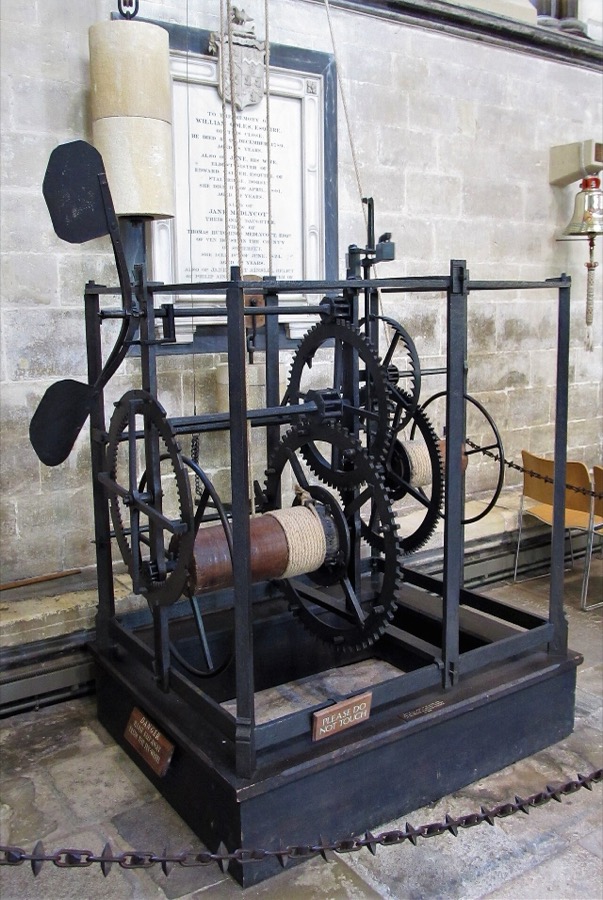 An open iron frame about waist high standing on a stone cathedral floor, holding two sets of large toothed wheels and a rope drum, with a horizontal bar carrying two flat weights above it, and nowhere on it a dial or a hand.