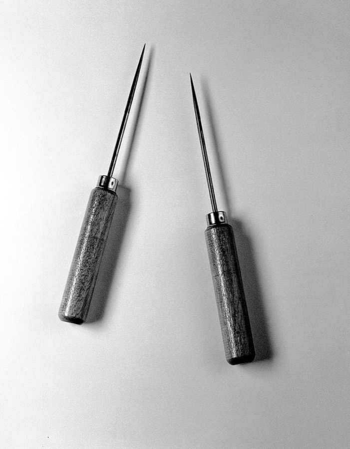 Black and white photograph of two slender surgical instruments on a pale background, each a plain wooden handle joined by a metal collar to a long steel shaft that tapers to a needle point.